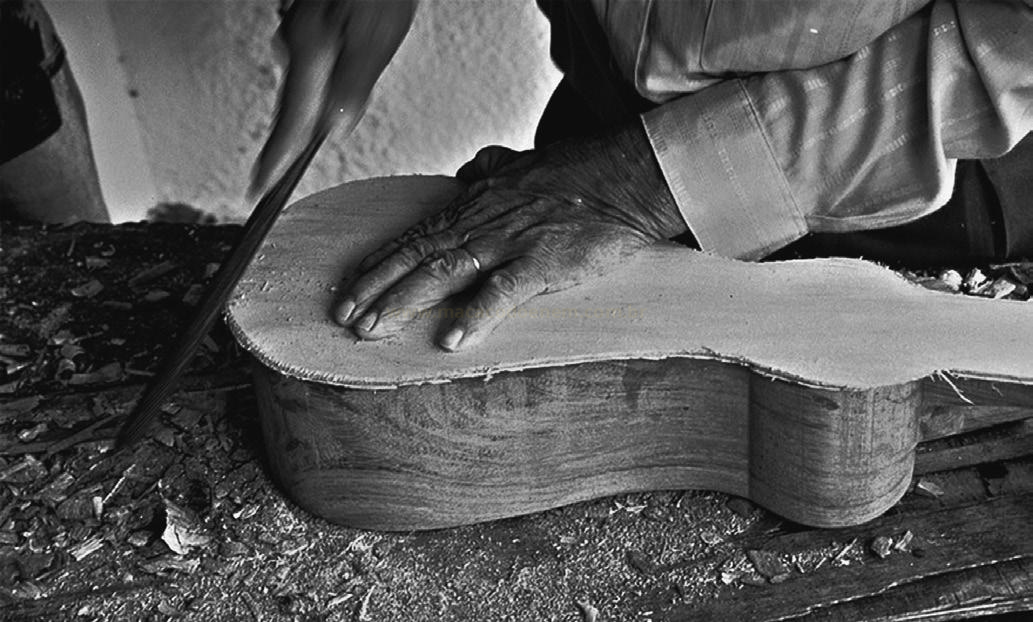 Fotografia em preto e branco, em close, de mãos trabalhando com formão no corpo de madeira de um violão.