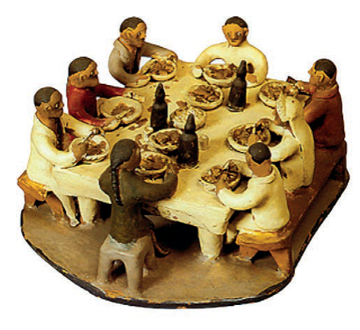 Escultura de cerâmica com várias pessoas sentadas ao redor de uma mesa posta, servindo-se de comida.