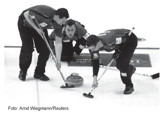 Fotografia em preto e branco de três jogadores de curling agachados, varrendo o gelo com vassouras diante de uma pedra.