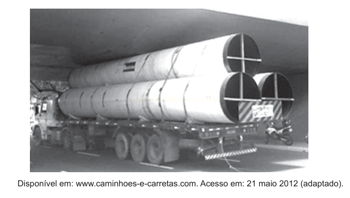 Fotografia em preto e branco de um caminhão carregando três grandes tubos cilíndricos sob um viaduto.