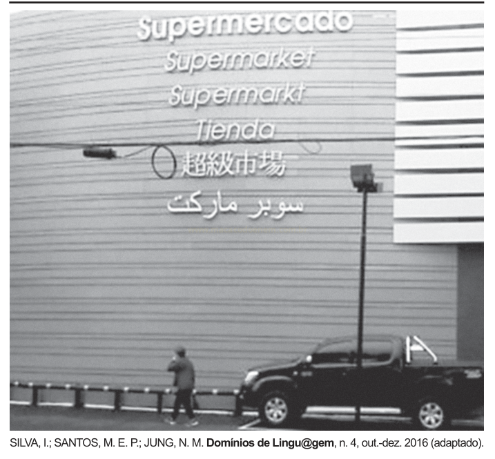 Fotografia em preto e branco de fachada com a palavra "supermercado" escrita em várias línguas, uma pessoa caminhando e uma caminhonete estacionada.