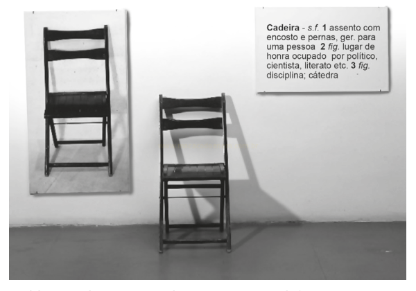 Fotografia em preto e branco com uma cadeira presa à parede, outra no chão e uma placa com o verbete 'Cadeira'.