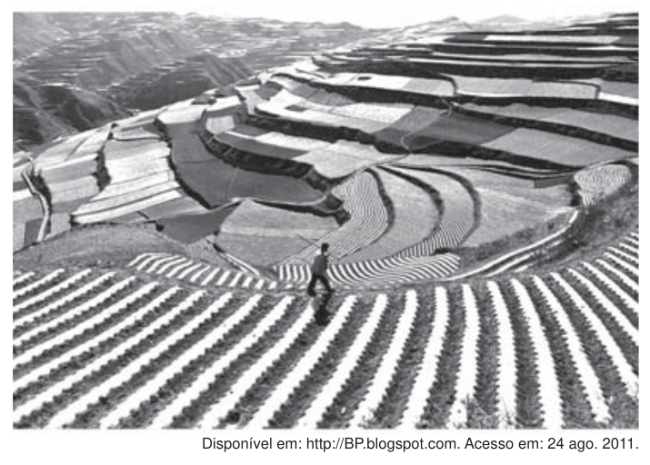 Fotografia em preto e branco de terraços agrícolas em encostas, com uma pessoa caminhando entre eles.