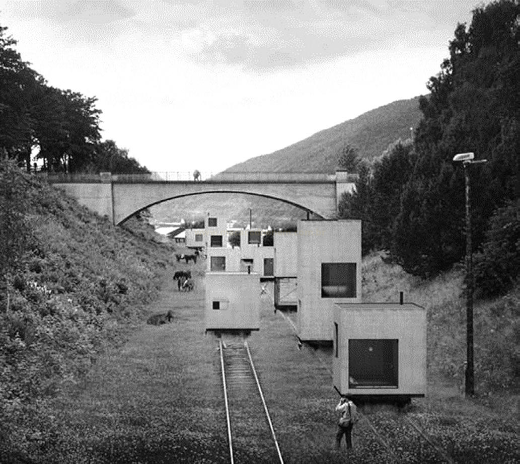 Fotografia em preto e branco de módulos cúbicos de concreto sobre trilhos de trem diante de uma ponte em arco.
