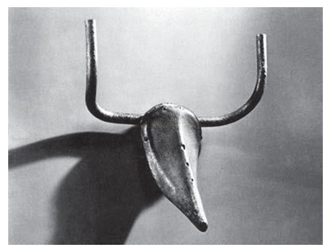 Fotografia em preto e branco de um objeto metálico com dois chifres longos e curvados, semelhante a uma cabeça de bovino, projetando sombra na parede.