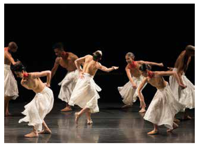 Fotografia de um grupo de bailarinos descalços com saias brancas em cena, sobre fundo escuro.