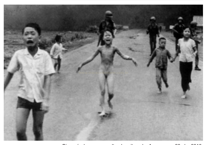 Fotografia em preto e branco de crianças correndo em uma estrada, com militares de capacete ao fundo.