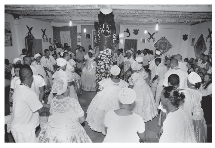 Fotografia em preto e branco de um grupo numeroso, vestido de branco com turbantes, reunido em círculo em torno de uma estrutura enfeitada com flores.