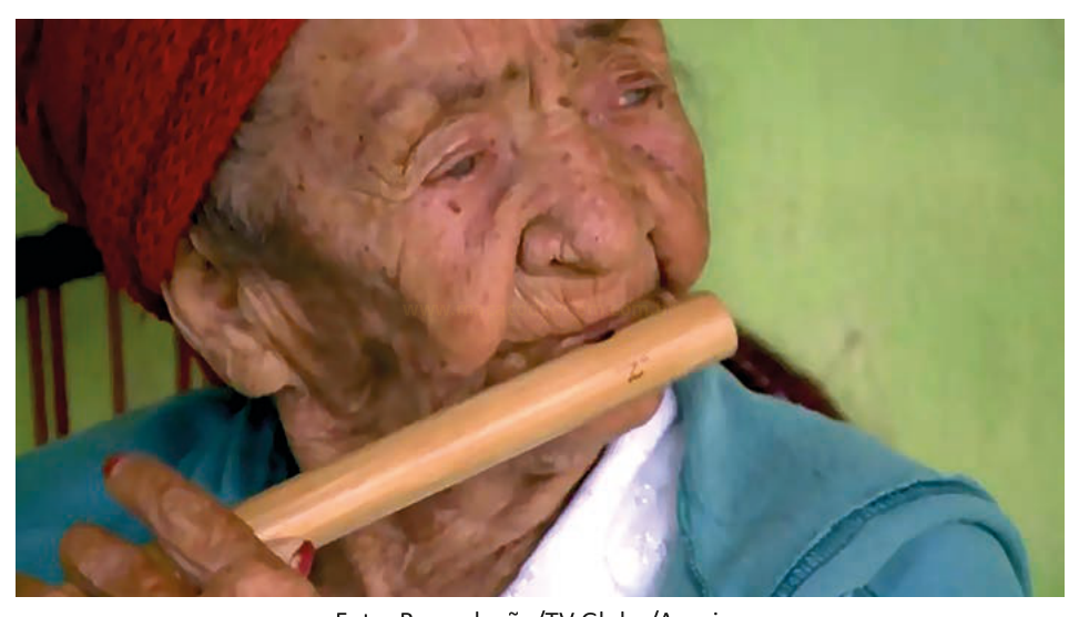 Fotografia de uma senhora idosa de gorro vermelho tocando uma flauta de bambu.