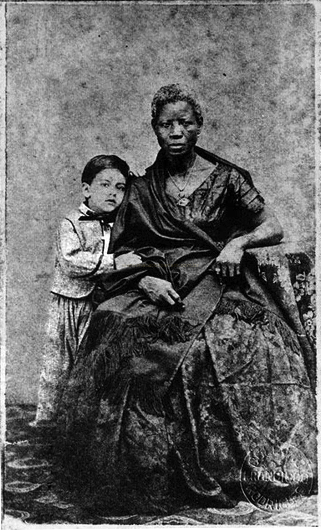 Fotografia antiga em preto e branco de uma mulher negra sentada com um menino apoiado ao seu lado.