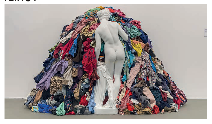 Fotografia de uma estátua branca clássica vista de costas diante de uma grande pilha colorida de roupas em uma galeria.