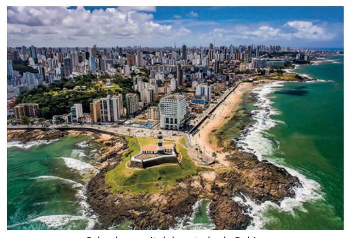 Vista aérea de cidade litorânea com farol em promontório rochoso, orla, mar e densa malha de edifícios ao fundo.