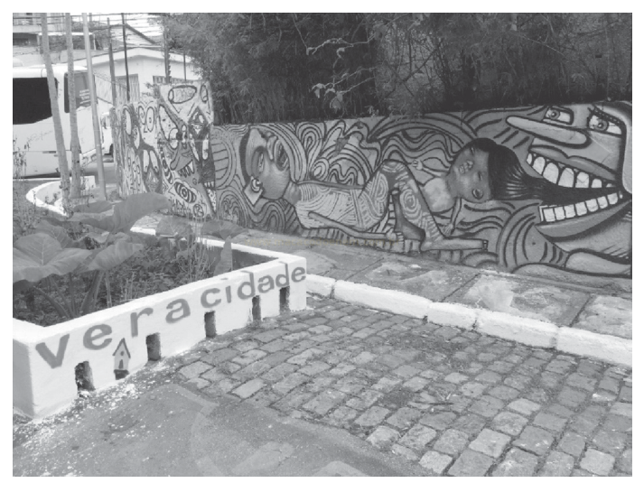Fotografia em preto e branco de um muro grafitado com figuras humanas estilizadas e a palavra "veracidade" em uma estrutura de concreto com degraus.