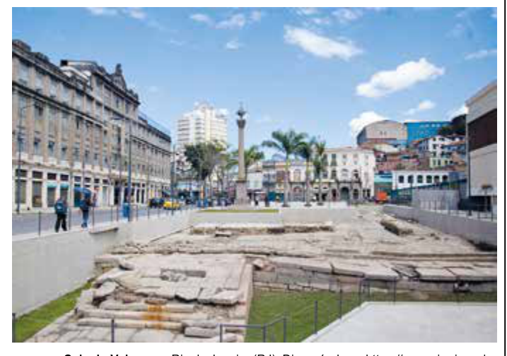Fotografia de uma praça urbana com ruínas arqueológicas cercadas em primeiro plano e edifícios antigos e modernos ao fundo.
