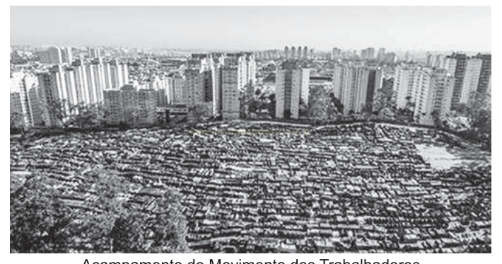Fotografia aérea em preto e branco de uma ocupação de casas baixas diante de edifícios altos ao fundo.