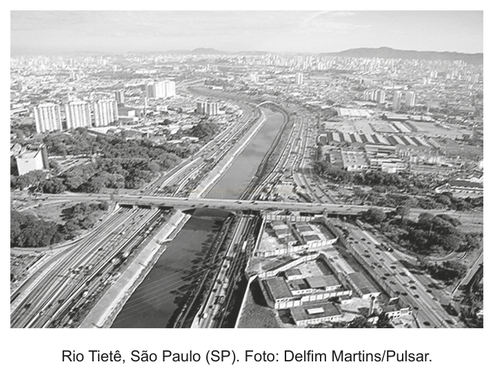 Fotografia aérea em preto e branco do rio Tietê em São Paulo, com uma ponte sobre o rio e a cidade ao redor.
