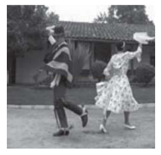 Fotografia em preto e branco de um casal dançando de costas, com um lenço erguido, em frente a uma casa.